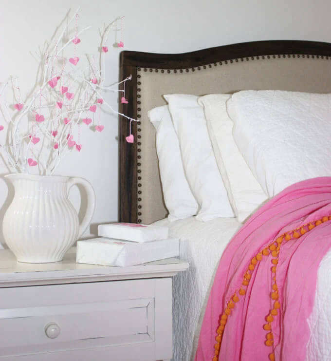 White vase on a white nightstand with white painted branches with min pink dangleing hearts.