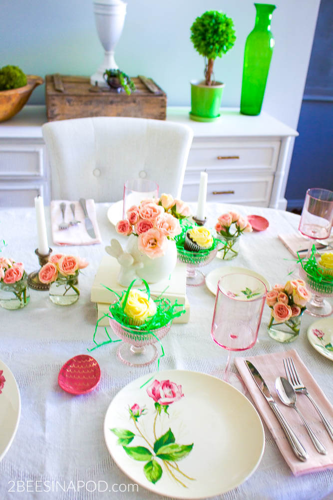 Easter Tablescape ideas. white tablecloth topped with pink rose stem plates, pink stemware, pink napkins, and pink bowls filled with easter grass and a cupcake.
