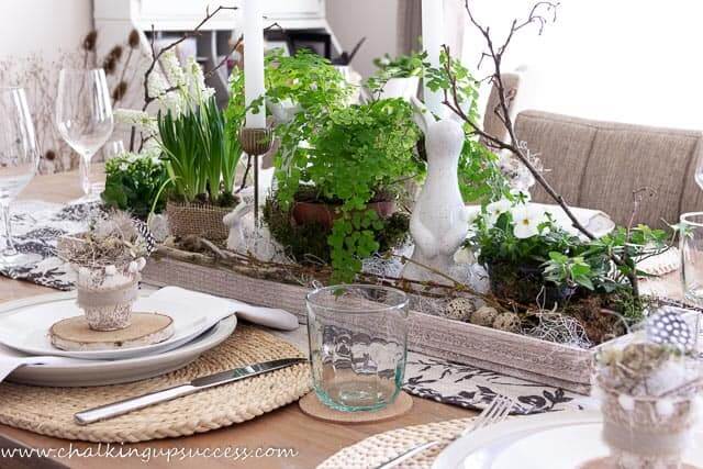 Natural woven placemats layered with white dishes and napkins. At each place setting you'll find a wood slice, clay pot filled with a nest and egg. Wooden tray is filled with green potted plants, candles, bunnies, eggs and twigs for the centerpiece.