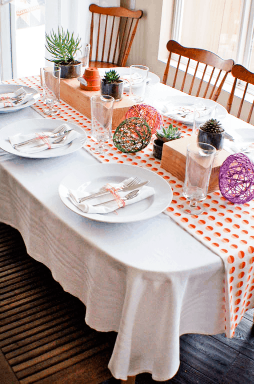 Orange and white polka dot tablerunner over white tablecloth. White plate with a white napkin, silverware, ties with an orange ribbon. Centerpiece consists of colorful string eggs and potted plants.