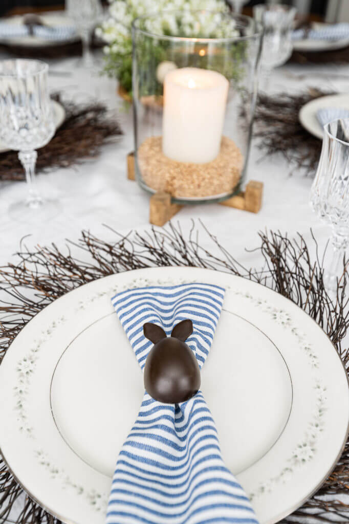 Easter place setting of white china plate place over a twig wreath topped with a blue and white striped napkin in a chocolate bunny napkin ring. Candle in a hurricane in the background.