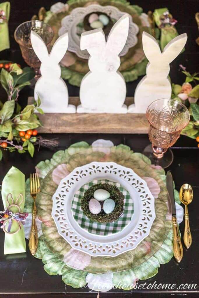 Easter Tablescape ideas, green petal placemat, layered with, amber and white dishes. Each place setting has a nest filled with eggs. Bunny shaped napkin rings with a green napkin. Wooden bunny centerpiece with florals.