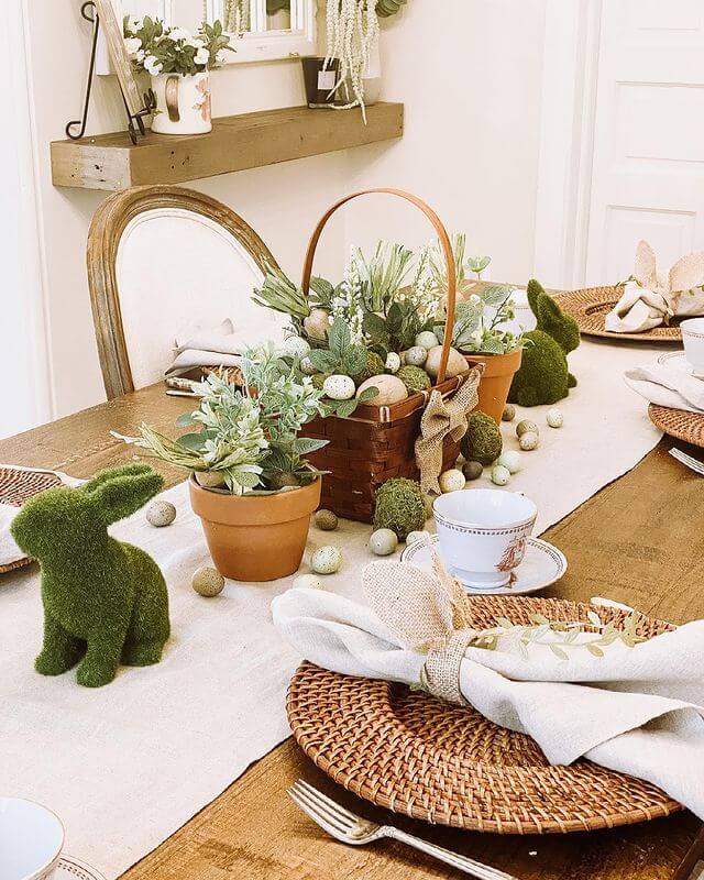 Easter Tablescape ideas, burlap tabletunner with moss bunnies, eggs and potted plants in clay pots. A Basket is placed in the center of the table filled with neutral carrots and eggs. At each place setting you'll find a natural rattan charger, linen napkin in a bunny ear napkin ring.