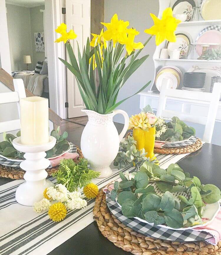 Yellow daffodils in white pitcher with candles, on a black and white table runner, natural grass placemats with black and white buffalo check chargers, white plates with a wreath.