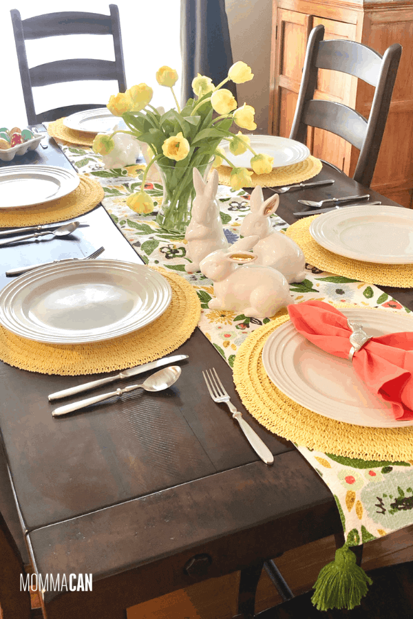 Easter Tablescape ideas, yellow and green table runner with a glass vase filled with yellow tulips, bunnies scattered down center of table. Yellow placemats wit white dishes.