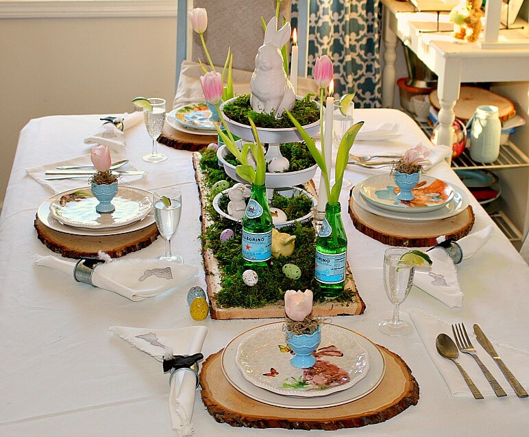 Rustic wood slices are used for chargers, layered with white and bunny plates. Centerpiece is a large wood slab layered with moss, eggs, bottles with tulip stems amd a tiered tray with bunnies and eggs.