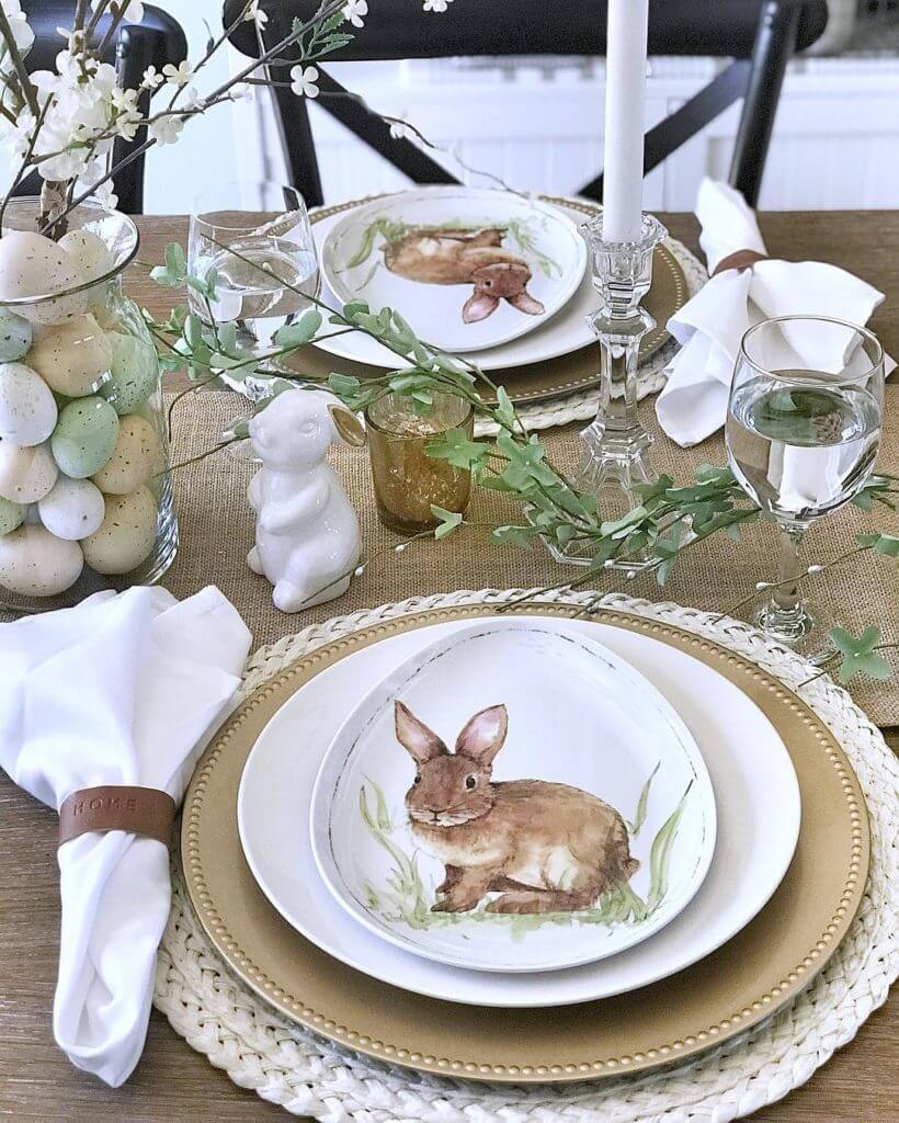 Burlap tablerunner, white woven placemats, gold charger, white plate with an egg shaped bunny plate. Spring blossoms in a glass vase filled with eggs.