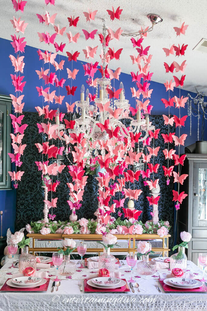 Easter Tablescape ideas, various shades of pink paper butterflies and strung from the ceiling over the dining table. Pink chargers, layered with white and pink dishes, a single pink flower is plated at each place setting in a bud vase.