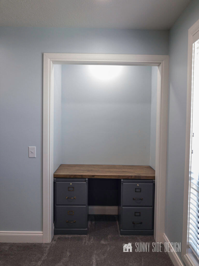 Cloffice area with grey painted wood file cabinets and natural wood floating desk top. Wall painted a light blue, with white trim.
