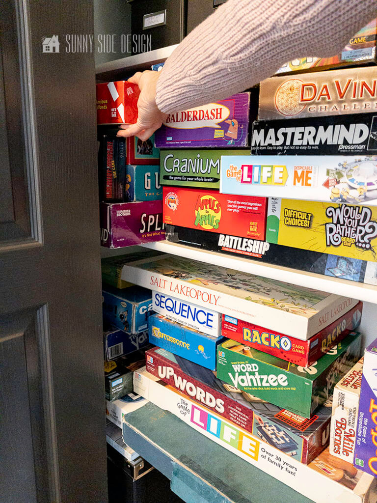 Woman's hand trying to remove a game from the corner of the closet. Many family board games stacked on the shelves in the closet.