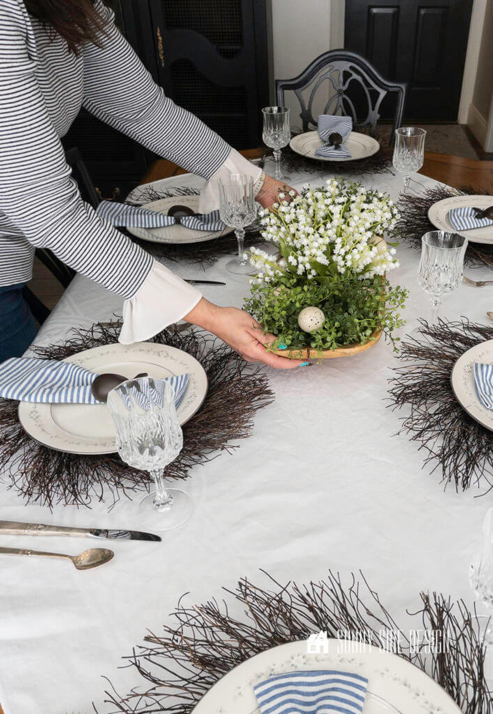Woman placing the centerpiec on the the Easter table decor. Wood bread bowl filled with greenery, Easter eggs, and white lily of the valley flowers.