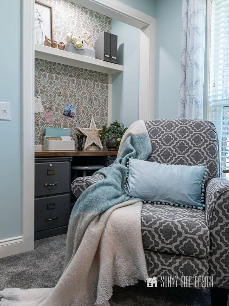 Cloffice, a small office in the closet space. Walls are a light blue with white trim and fabric with a grey and blue pattern has been applied above the desk for a pin board and above the shelf. Grey patterned chair is in front of the cloffice area.