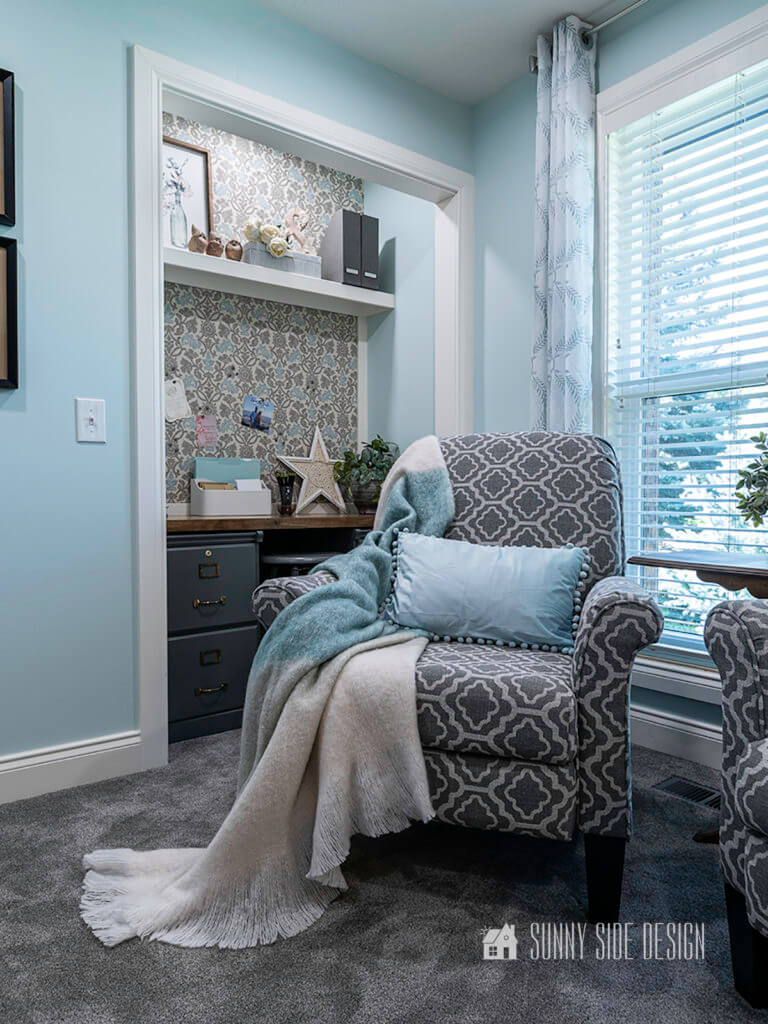 Cloffice, a small office in the closet space. Walls are a light blue with white trim and fabric with a grey and blue pattern has been applied above the desk for a pin board and above the shelf. Grey patterned chair is in front of the cloffice area. White and blue patterned sheers frame the window.