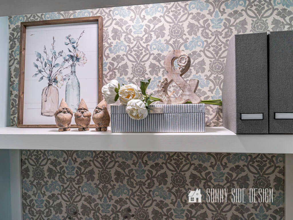 Floating shelf above desk area, staged with framed art, fabric boxes for storage, flowers, grey fabric magazine boxes and 3 wooden gnomes.
