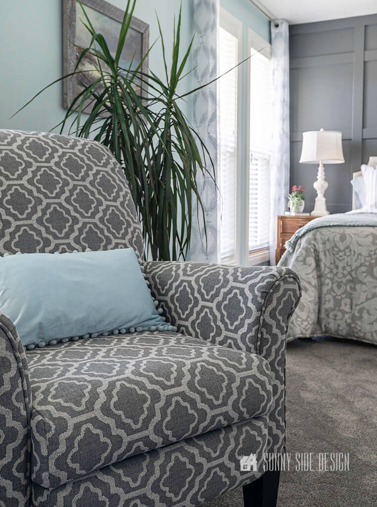 Gray and white patterned fabric chair in forground. Light blue walls with oil painting of seascape and dark gray board and batten wall in background with nightstand and bed.