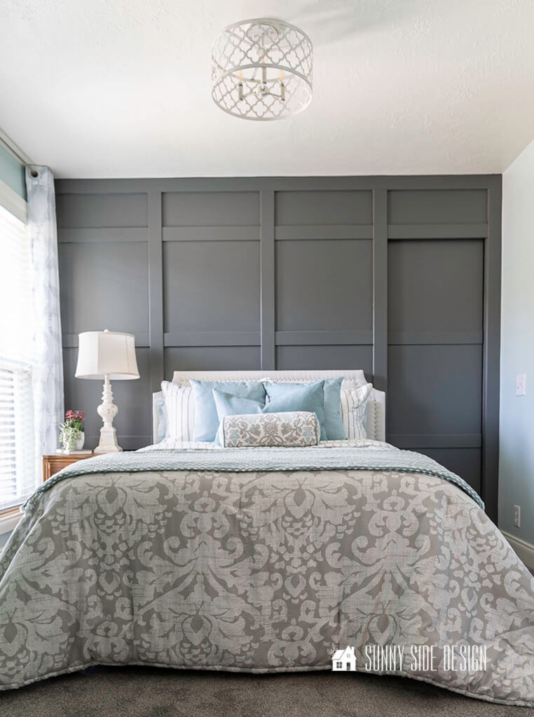Dark gray board and batten wall with french style wood nightstand, white distressed lamp, white upholstered bed with nailhead trim., blue pillows and gray and white damask bedding.