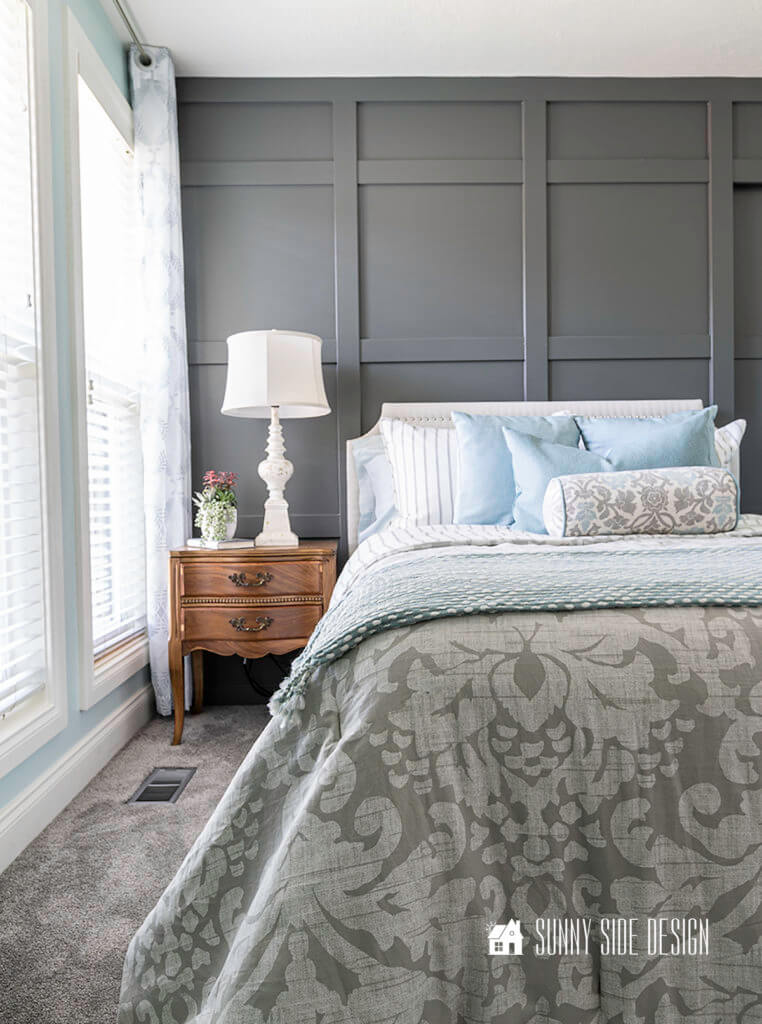 Dark gray board and batten wall with french style wood nightstand, white distressed lamp, white upholstered bed with nailhead trim., blue pillows and gray and white damask bedding.