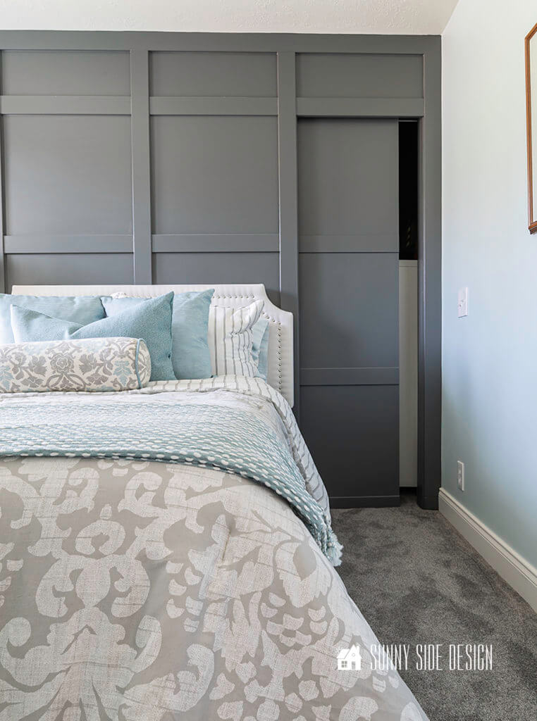 Dark gray board and batten wall with pocket door. Surrounding walls light blue. White upholstered headboard with nailhead trim against dark gray wall, gray and white damask beddint with blue addent pillows.