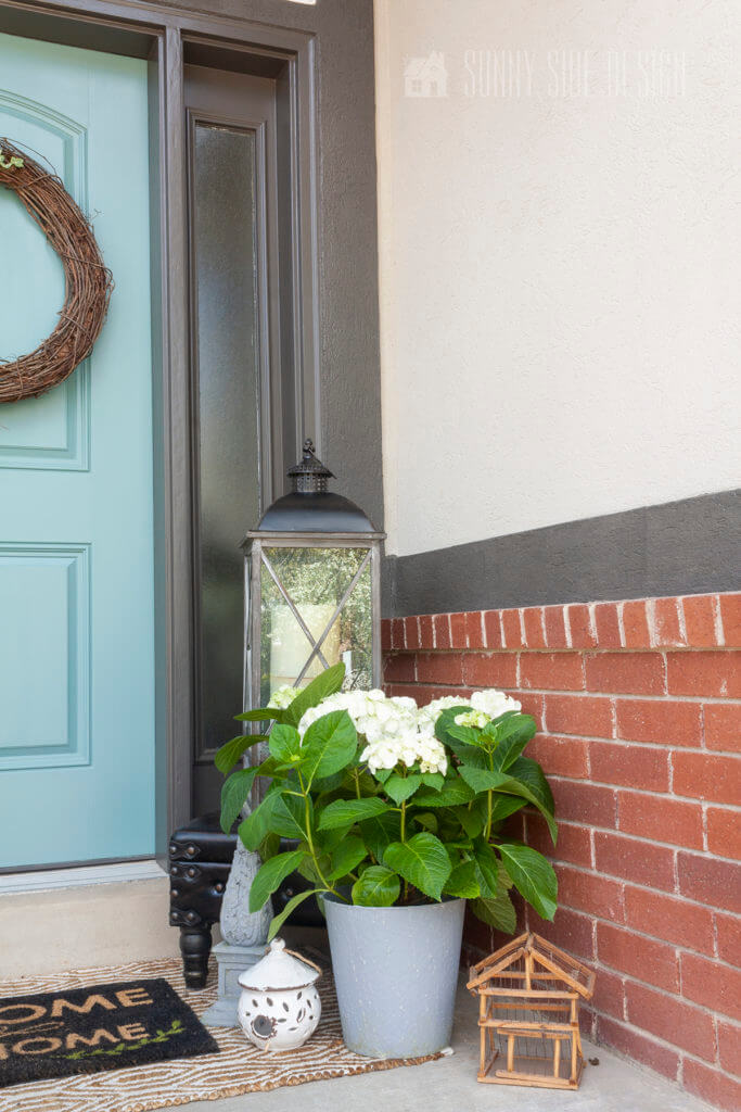 Curb appeal ideas, Potted white hydrangeo with a large lantern, and birdhouses. Red brick and taupe stucco home with a dark charcoal grey trim, blue front door.
