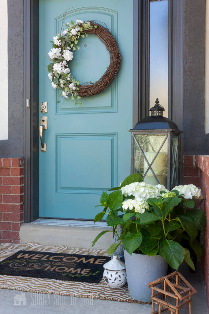 Curb appeal ideas, Potted white hydrangeo with a large lantern, and birdhouses. Red brick and taupe stucco home with a dark charcoal grey trim, blue front door with a grapvine and white geranium wreath.