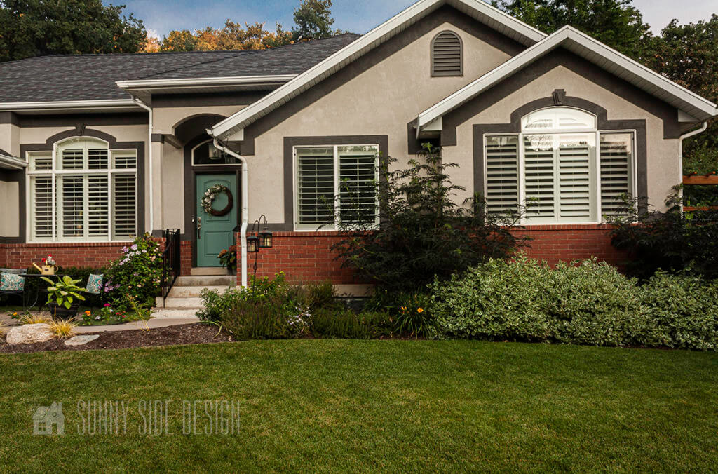 Curb appeal ideas, Red brick home with taupe stucco with the pop out painted dark charcoal grey. The front door is painted a slate blue. The landscaping has been updated. Seating area near the front door.