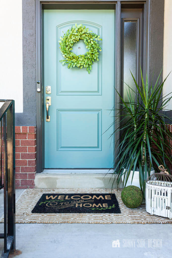 Curb appeal ideas, slate blue door with dark charcoal grey trim, taupe colored stucco, red brick. Green wreath on door, welcome mat layered with sisal rug, potten plant and birdcage.
