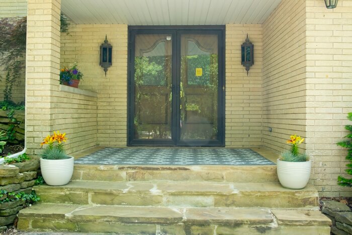 White brick home with brown double doors with a glass security door, dated lighting, potted flowers on steps and a large blue rug.