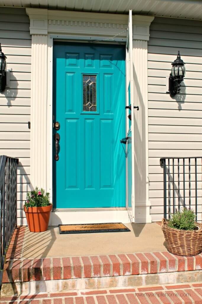 Curb appeal ideas, newly painted front door a beautiful teal color, white trim surround door, grey clapboard sidding, and a brick porch with potted flowers.