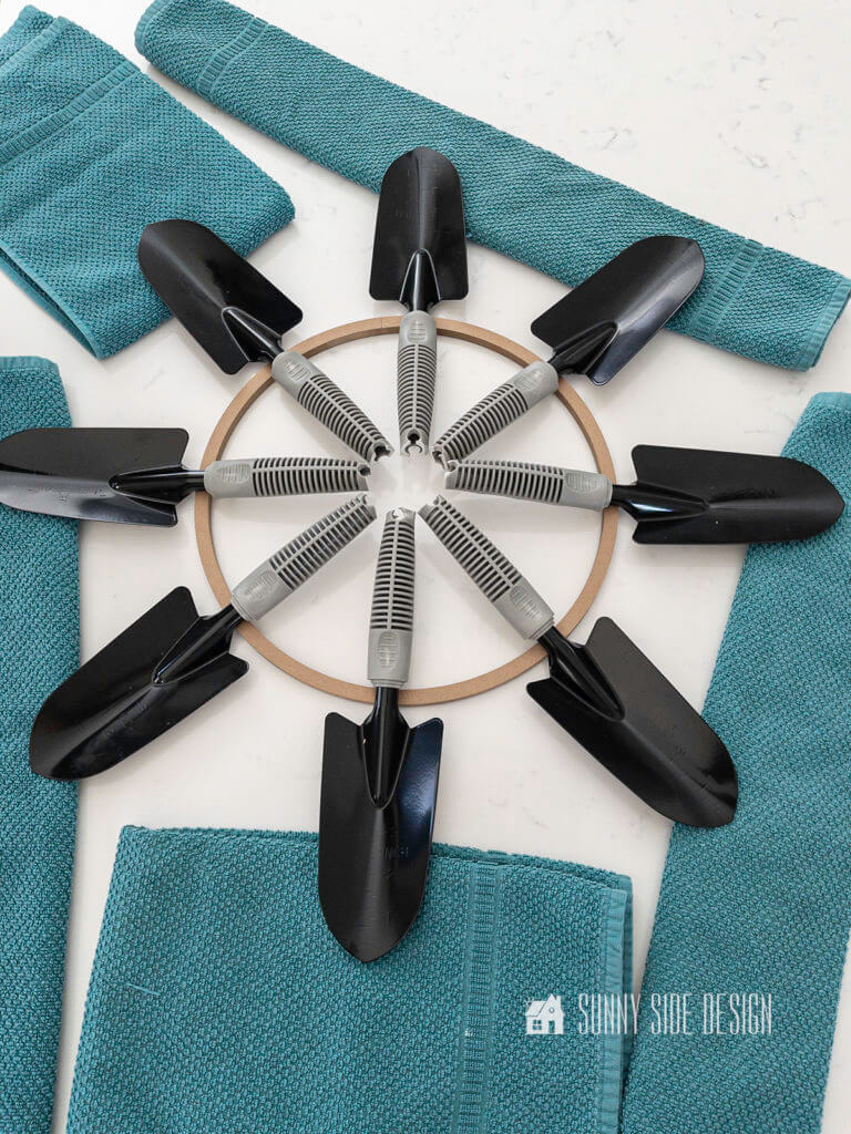 Eight black trowels are placed on top of the wood floral ring evenly spaced forming the summer garden wreath. Towels are used to keep trowels level.