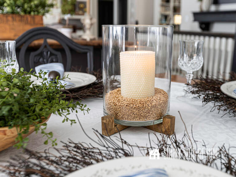 Pottery Barn dupe, lit candle in a glass hurricane on a wood stand setting on an Easter decorated table.