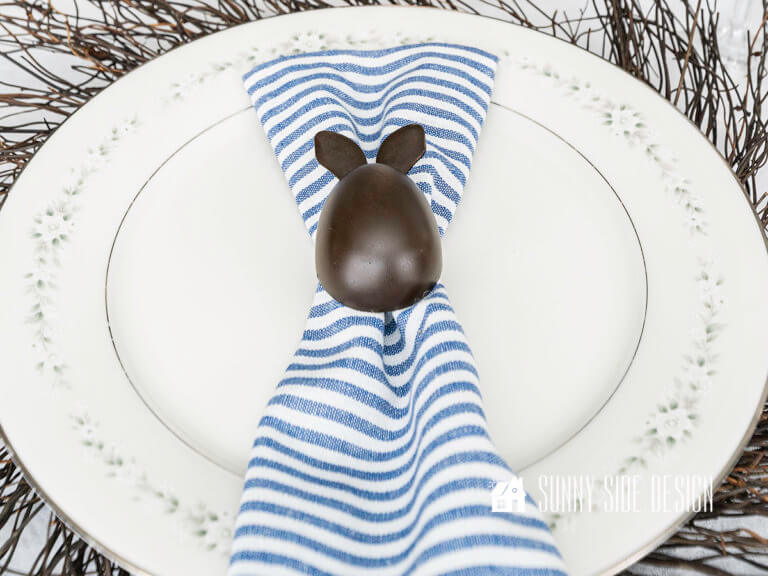 Faux Chocolate bunny Easter egg on a blue and white striped napkin, placed on a white with silver rim china plate on a twig wreath.