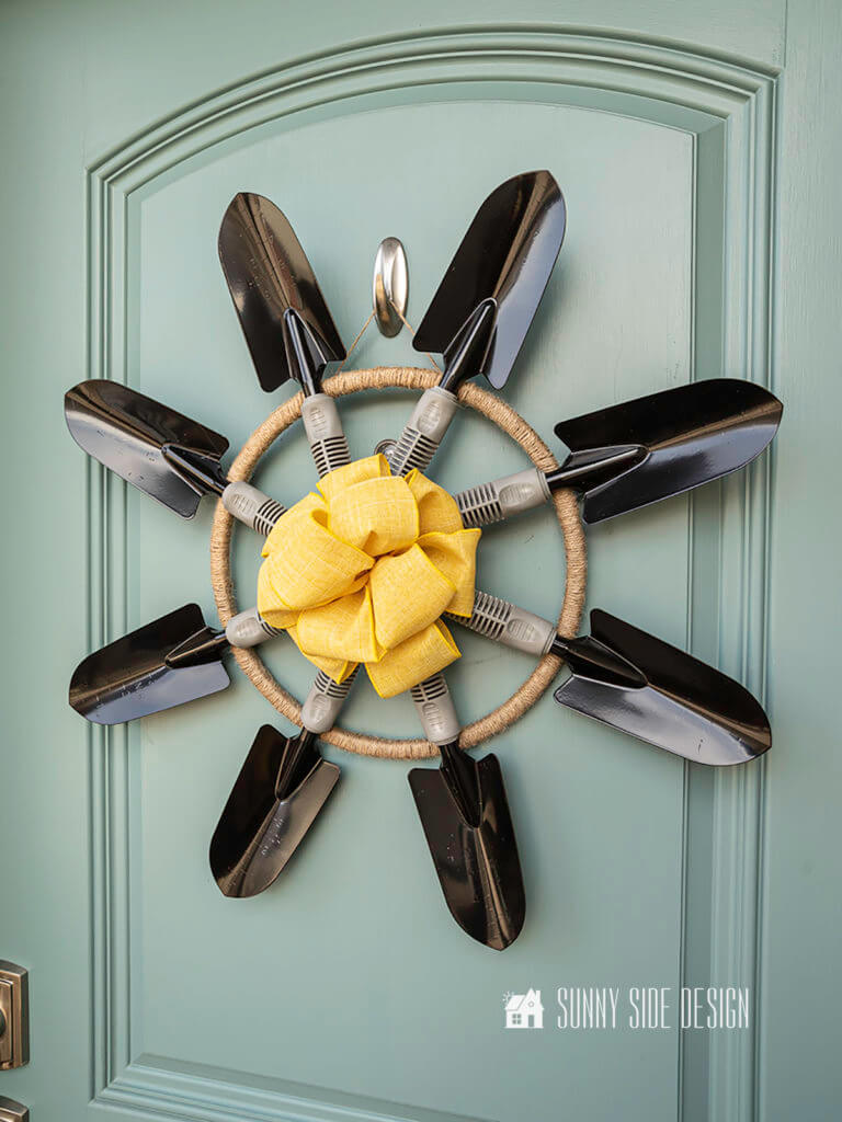 Eight black garden trowels forming a unique summer garden wreath with a yellow bow in the center, hanging on a blue door.