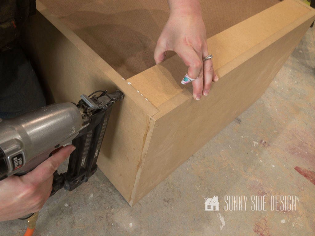Woman holds brad nailer against cabinet side to attach the back board to make a cabinet.