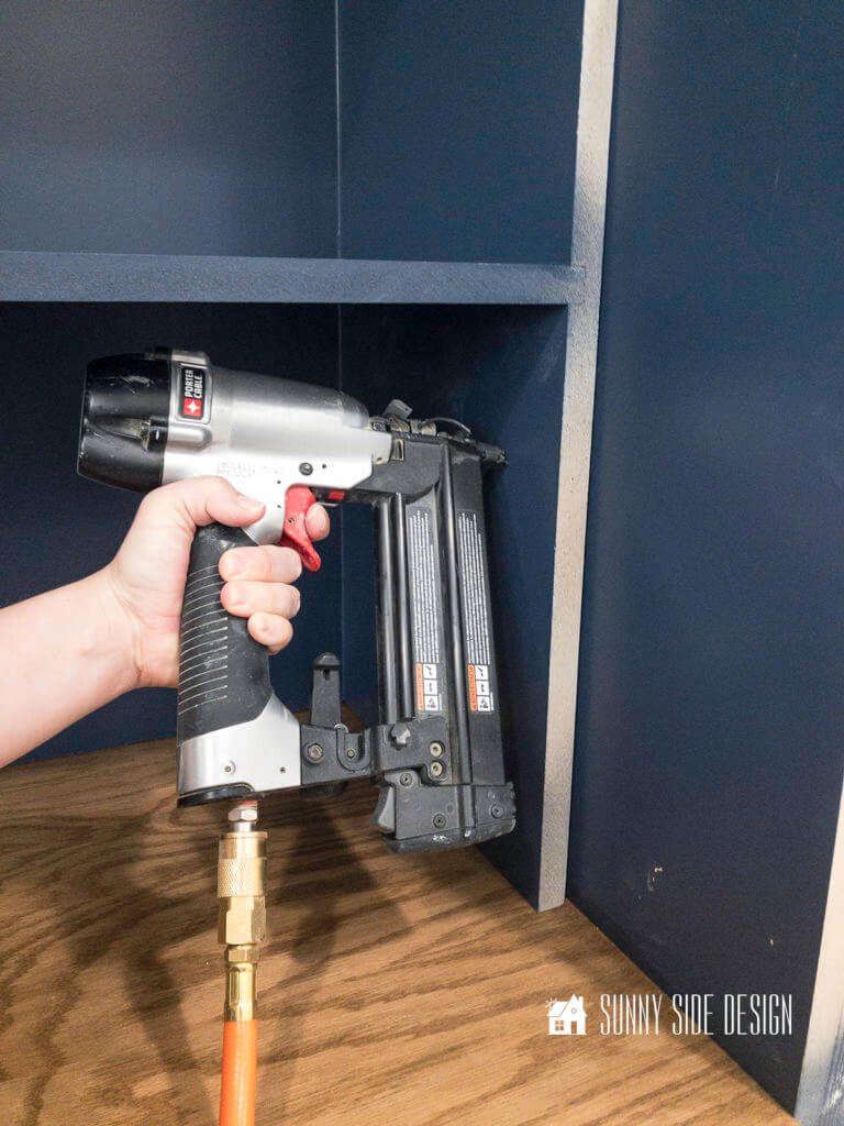 Woman's hand holding a brad nailer, attaching the booksleves to the side cabinet.