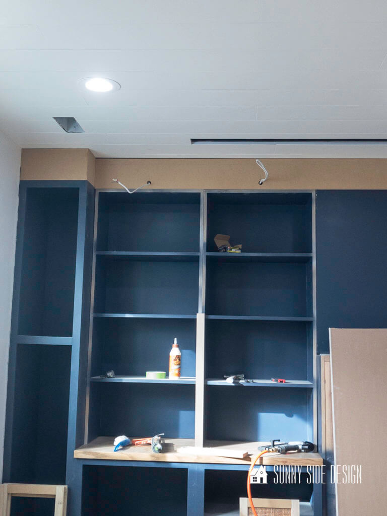 Wood is installed along the top of the built in entertainment center to the ceiling with wire coming through holes for the library lighting.