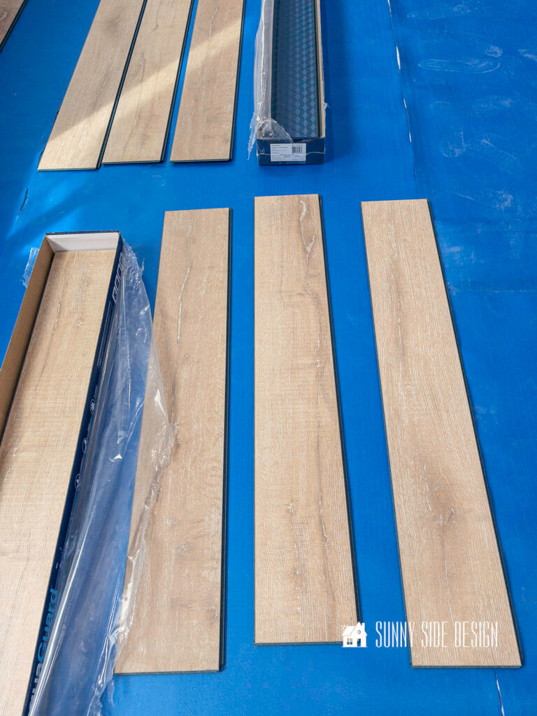 Several pieces of the laminate flooring are layed out from different boxes, mixing patterns and colors on the blue waterproof underlayment.