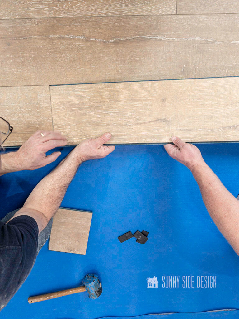 Man's hands tilting laminate flooring to lock short end of laminate into previous piece, blue waterproof underlayment.