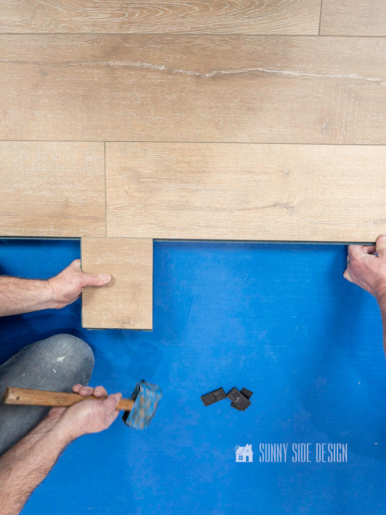 Man hands holding a rubber mallet and a scrap piece of laminate flooring, tapping laminate into place.