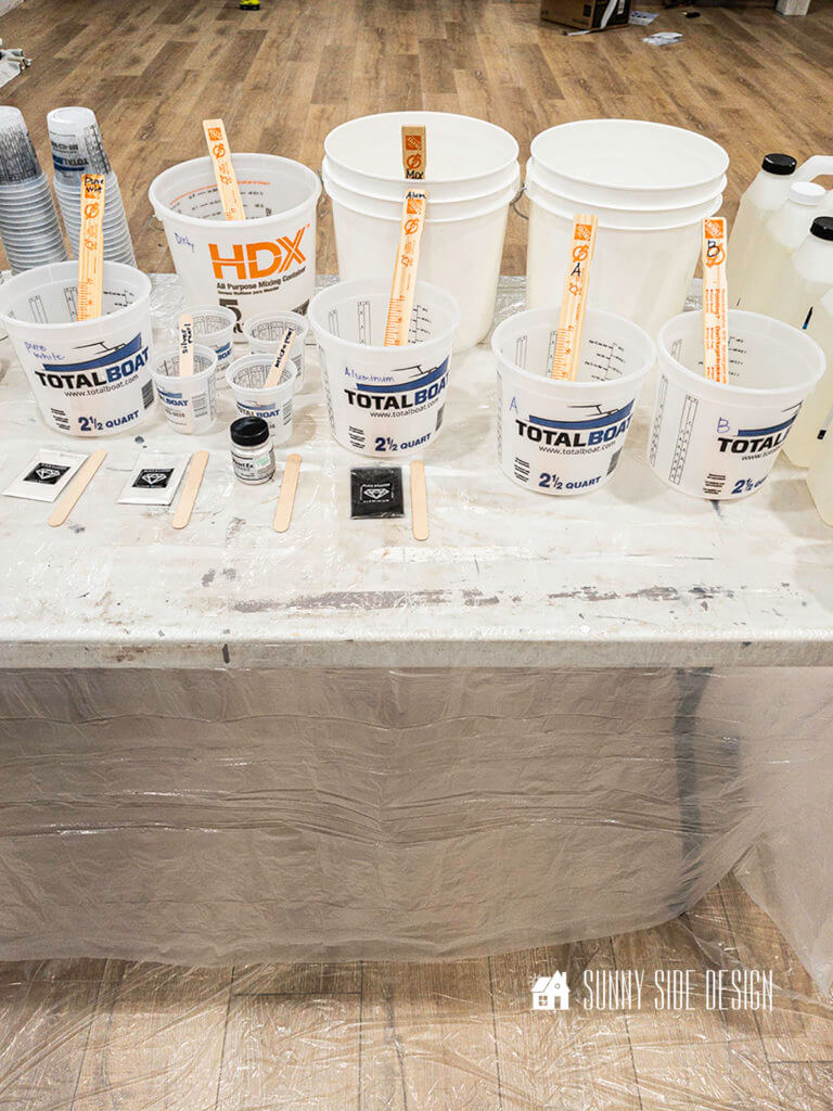 Buckets are lined up on a table covered with plastic with paint stirsticked marked with the color of the epoxy product.