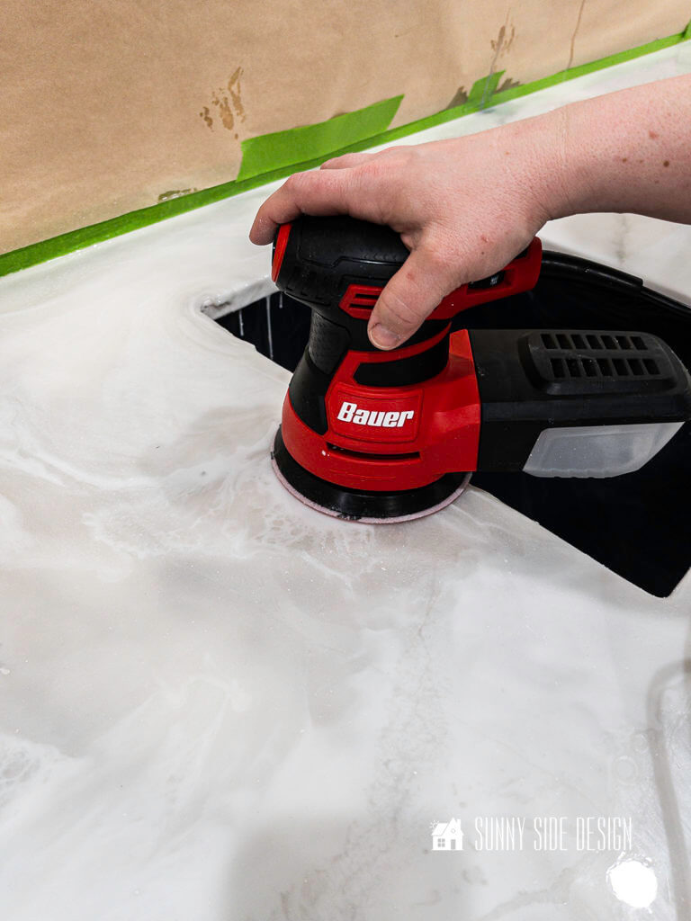 A woman's hand holding a orbial sander, sanding the edge of the epoxy countertop.