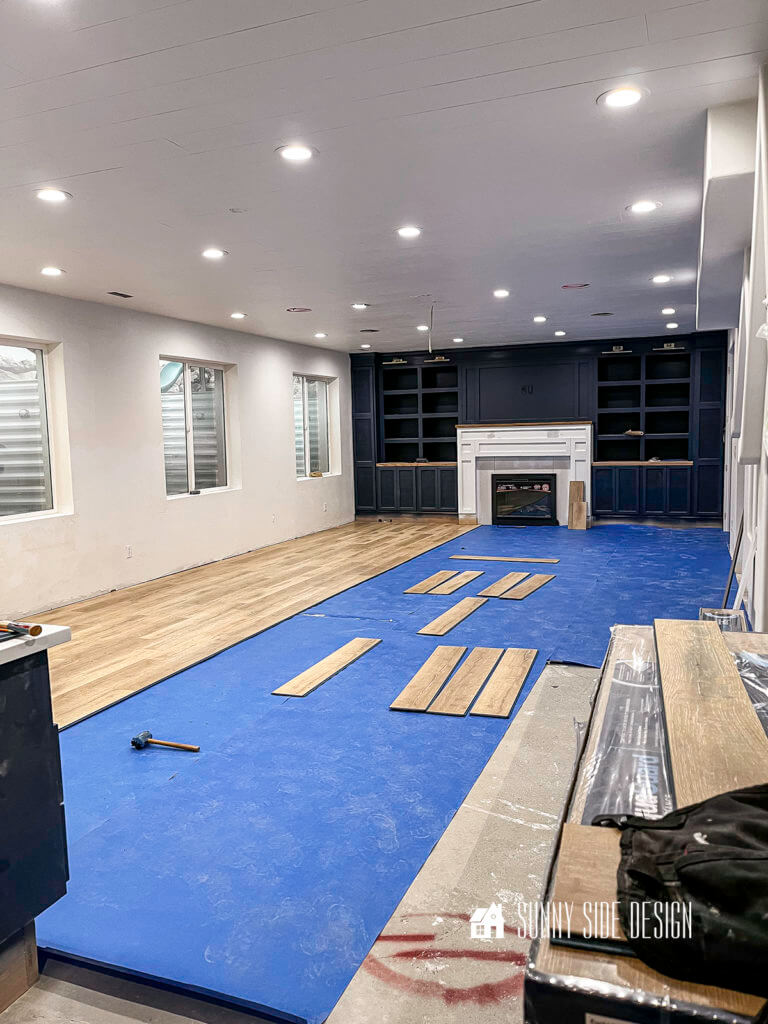 Install a laminate floor over concrete about 1/3 of the laminate is installed in the family room, with blue waterproof layer covering the remainder of the floor, Navy blue built in entertainment wall with a white fireplace surround.