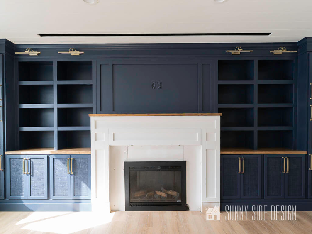 Full view of built in entertainment center with Hale Navy blue painted cabinets and shelves with rattan doors and champagne gold hardware. Center of the built in is a white fireplace mantle and surround with an oak mantle.
