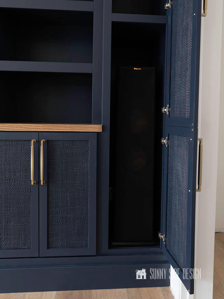 Right side of built in entertainment center with Hale Navy blue painted cabinets and shelves with rattan doors and champagne gold hardware. Door open showing large speaker that is concealed behind the rattan doors.