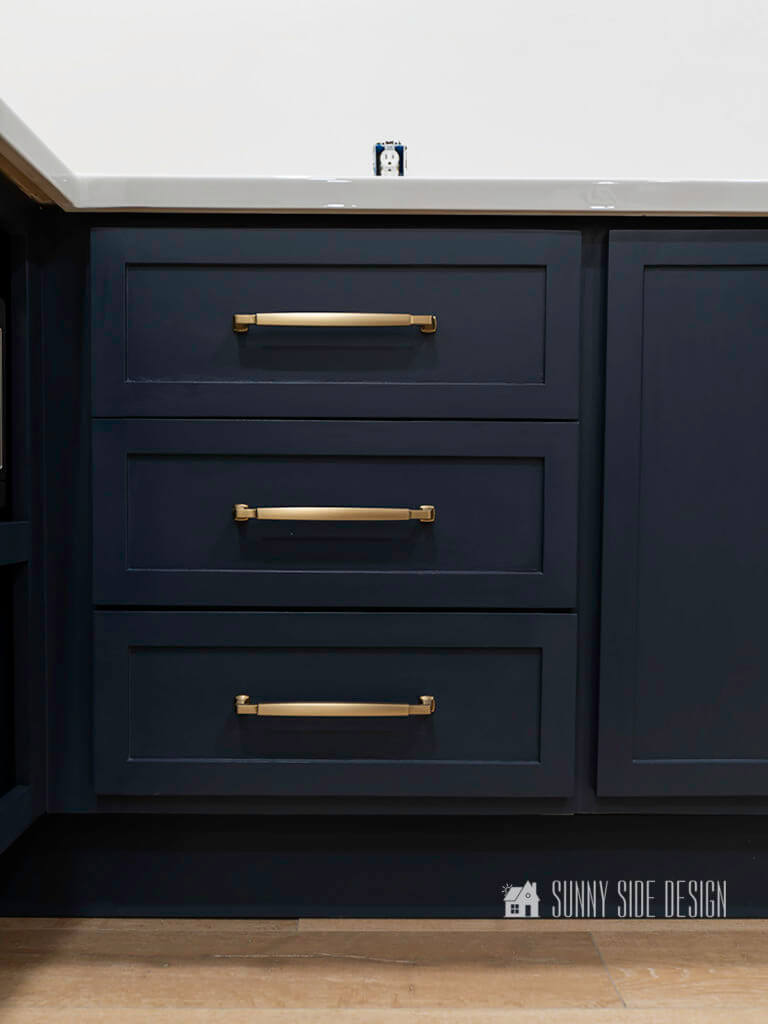 Navy blue shaker style base kitchen cabinets with champagne gold drawer pulls and a natural oak flooring with white walls.