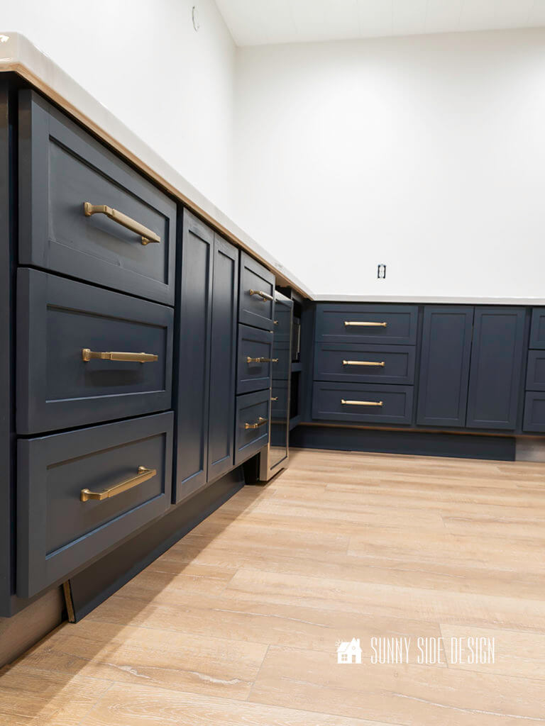 Navy blue shaker style base kitchen cabinets with champagne gold drawer pulls and a natural oak flooring with white walls.