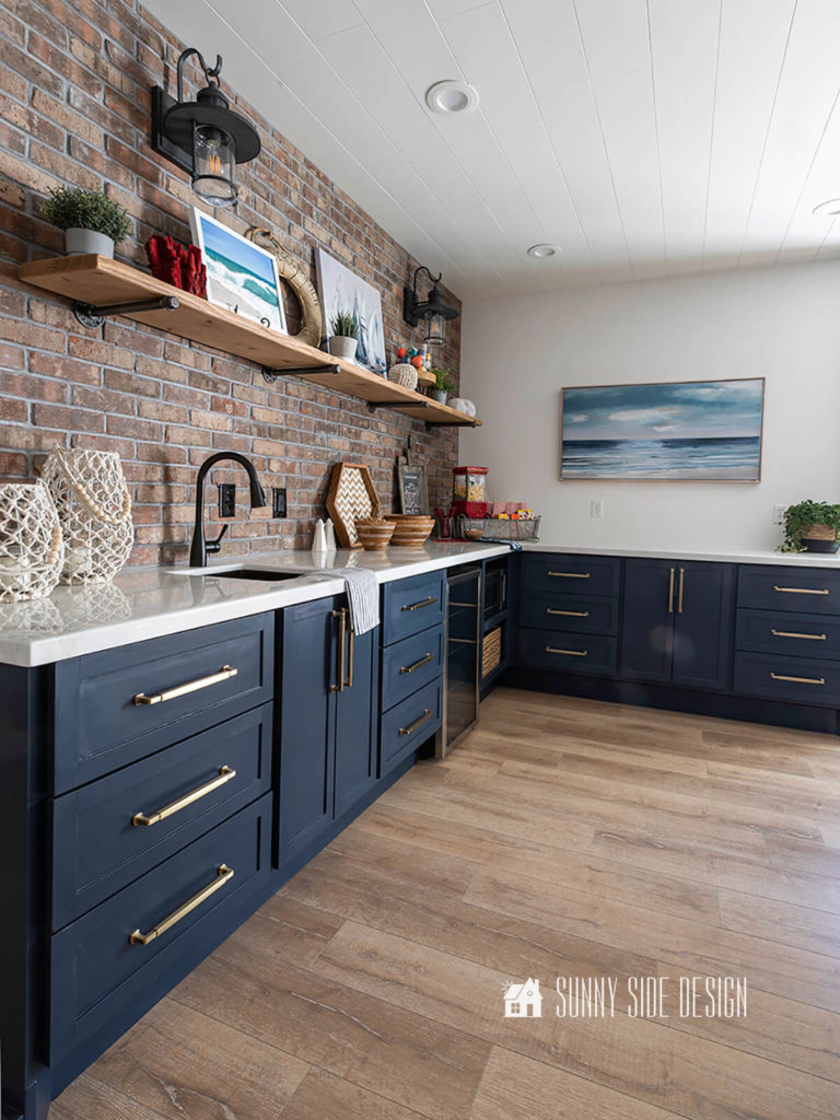 Navy blue kitchen cabinets with champagne gold hardware and a rustic brick backsplash, marble look epoxy countertops. Industrial pipe and wood shelf decorated with art and accessories.