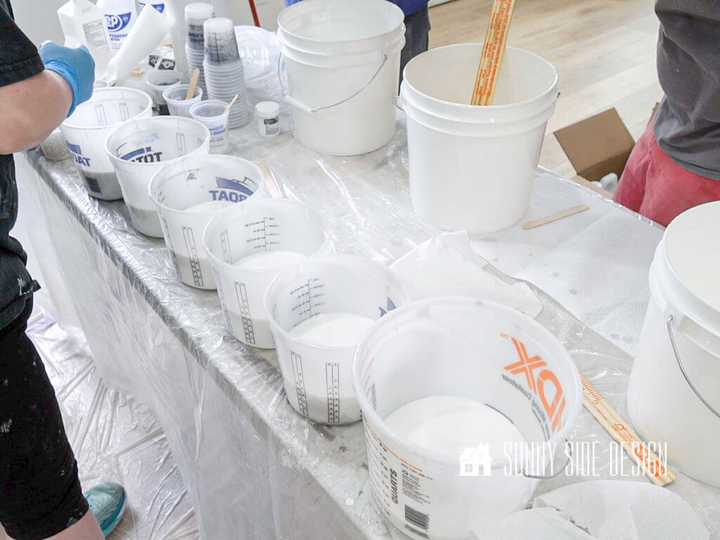 6 buckets are lined up with epoxy in each bucket on a table covered with plastic.