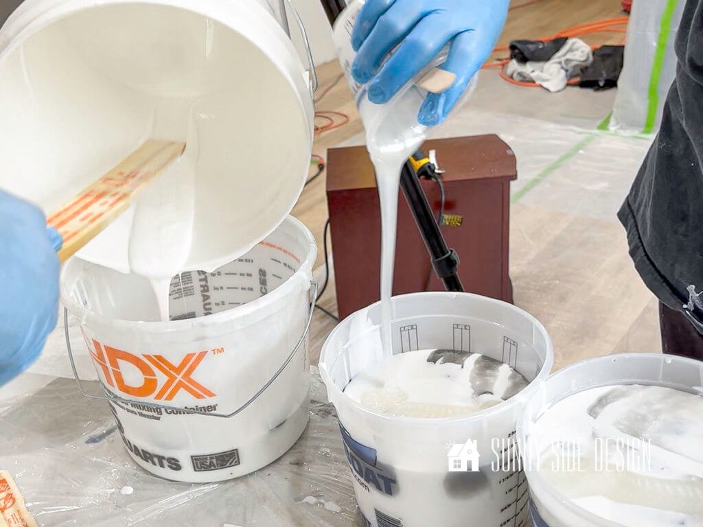 Woman's hand wearing blue latex gloves is pouring white pearl epoxy into a bucket of black and white epoxy.