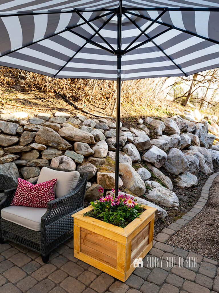 Mobile planter box with umbrella stand on a paver patio styled with a wicker chair and pink pillow