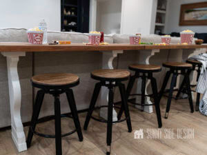 DIY SOFA TABLE WITH BLACK METAL AND WOOD SEAT BAR STOOLS. STAGED WITH POPCORN, CANDY AND BEVERAGES.
